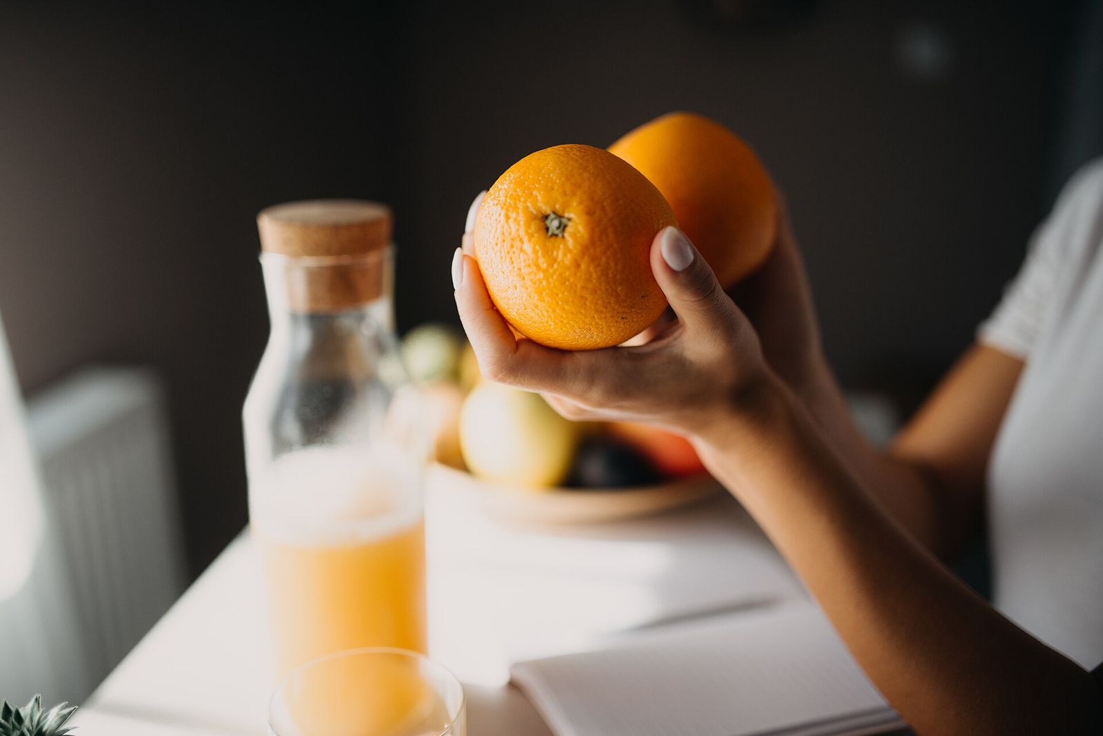 A hand lifting an orange from the fruit bowl on a sunlit breakfast table, beside a jug of juice and an open notebook